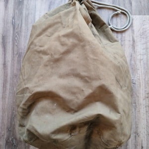 May include: A tan canvas drawstring bag with a rounded shape. The bag has a light-colored drawstring closure and a textured appearance. The bag is likely used for storage or carrying items.