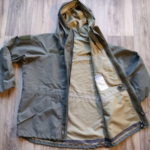 May include: An olive green hooded jacket with a full-length zipper, open to reveal a lighter interior. The jacket has multiple pockets and a drawstring waist. The hood is folded back, and the jacket is laid flat on a wooden surface.