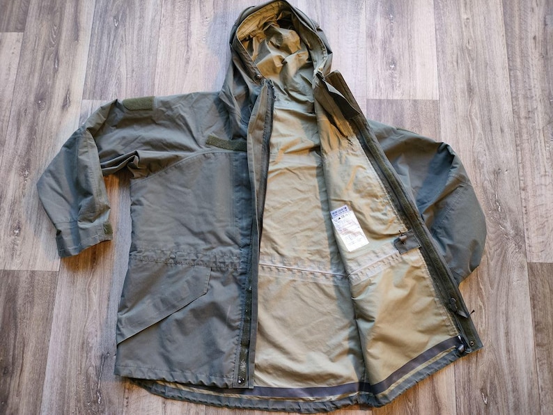 May include: An olive green hooded jacket with a full-length zipper, open to reveal a lighter interior. The jacket has multiple pockets and a drawstring waist. The hood is folded back, and the jacket is laid flat on a wooden surface.