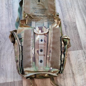 Puede incluir: Una mochila de campo de camuflaje con una solapa grande que se abre para revelar un compartimento con cremallera. La solapa tiene el texto "RCT SHAKLEY 305-021-18 FIELD PACK 8465-99-434-6734" impreso en ella.