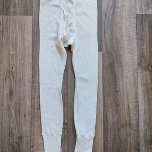 Peut inclure: Bas de sous-vêtements thermiques crème avec une texture en nid d'abeille. Le vêtement a une braguette boutonnée et des étriers aux chevilles. La ceinture est d'une nuance plus claire de crème.