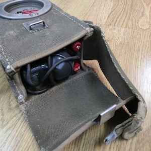 May include: A green canvas bag with a metal clasp and a black cord. The bag is open and contains a black and gray device with a red knob and a black cord. The bag has a strap.