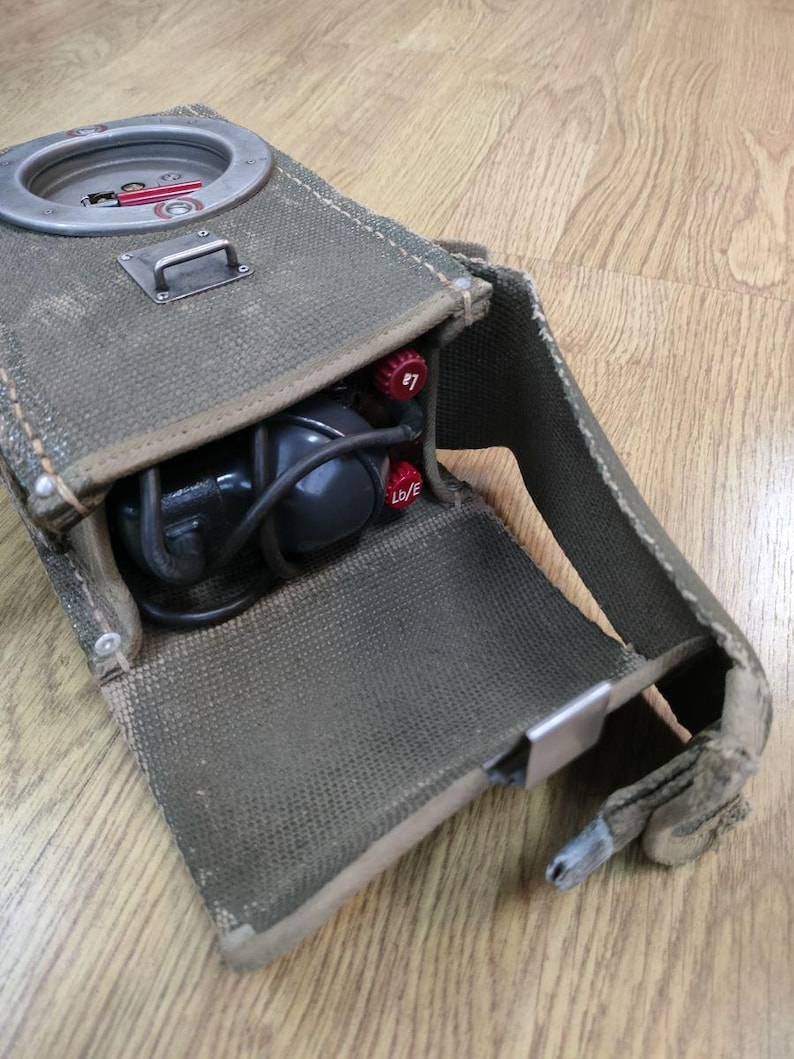 May include: A green canvas bag with a metal clasp and a black cord. The bag is open and contains a black and gray device with a red knob and a black cord. The bag has a strap.