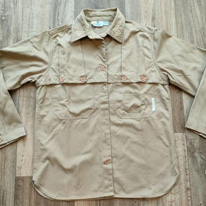Pode incluir: Uma camisa bege de manga comprida com botões, bolso com aba e colarinho abotoado.