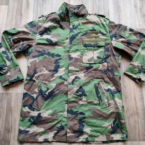Può includere: Giacca mimetica nei toni del verde, marrone e blu navy. La giacca presenta un colletto alto, tasche multiple e una chiusura con bottoni. Le maniche hanno polsini con bottoni. La giacca è appoggiata su una superficie di legno.