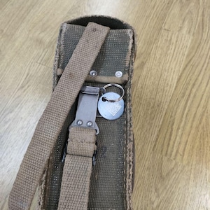 May include: A green canvas bag with a brown strap and a metal buckle. The bag has a key ring attached to it.