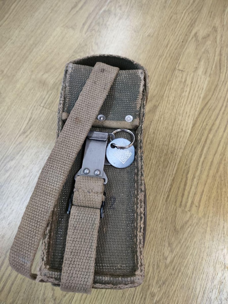 May include: A green canvas bag with a brown strap and a metal buckle. The bag has a key ring attached to it.