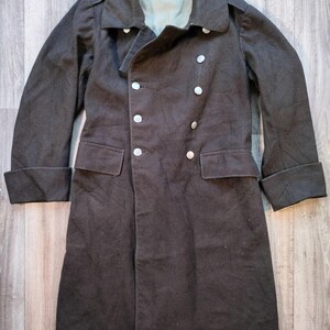 Può includere: Un cappotto marrone scuro a doppio petto con grandi bottoni color argento. Il cappotto ha un ampio colletto, due tasche anteriori e polsini ripiegati. La fodera interna è di colore verde chiaro. Il cappotto è esposto su una superficie di legno.