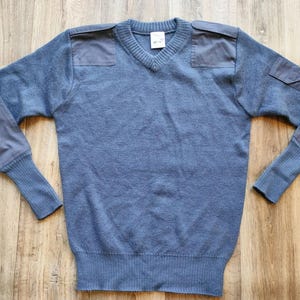 Puede incluir: Un suéter azul con cuello en V con parches reforzados en los hombros y los codos. El suéter tiene puños y dobladillo acanalados. La prenda está hecha de un material suave y tejido. El suéter se muestra sobre una superficie de madera.