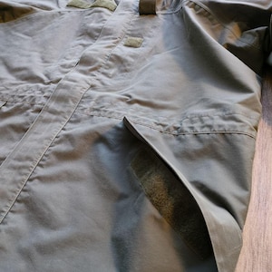 May include: Olive green hooded jacket with multiple pockets and velcro patches. The jacket has a full zipper closure and a high collar. The fabric appears to be a durable, weather-resistant material, suitable for outdoor activities. The jacket is designed for functionality and protection.