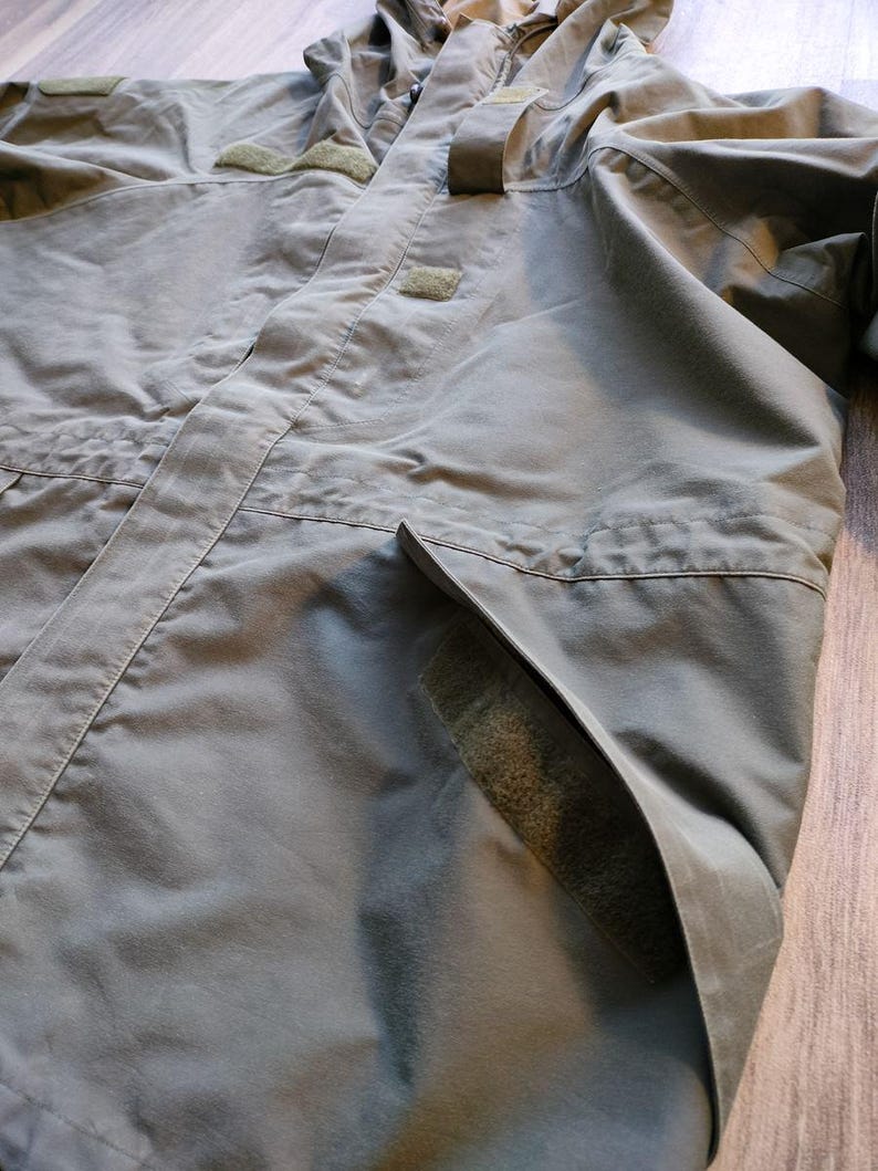 May include: Olive green hooded jacket with multiple pockets and velcro patches. The jacket has a full zipper closure and a high collar. The fabric appears to be a durable, weather-resistant material, suitable for outdoor activities. The jacket is designed for functionality and protection.