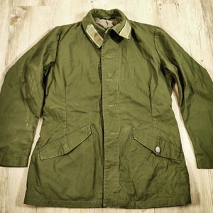 Può includere: Una giacca militare verde con chiusura a bottoni e due tasche a patta. La giacca ha un colletto con un motivo mimetico verde e marrone.