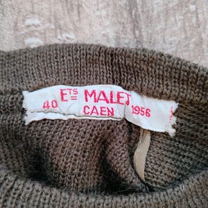 Puede incluir: Una etiqueta blanca con texto negro que dice "Ets Malet Caen 1956" y el n&uacute;mero "40" en una tela de punto marr&oacute;n.