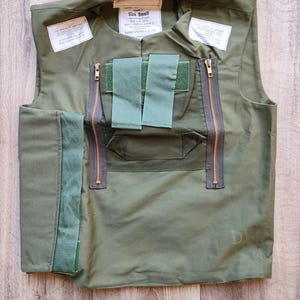 Op de afbeelding: Olijfgroen vest met twee verticale ritsen, twee groene stoffen banden en meerdere witte labels. Het vest is gemarkeerd met "Size Small". Het vest is gemaakt van een duurzame stof en heeft een militair design.