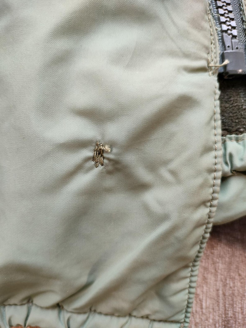 Może przedstawiać: Zbliżenie na zieloną nylonową kurtkę z zamkiem błyskawicznym i małą dziurką w materiale.