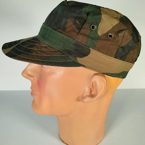 May include: A camouflage green, brown, and black military style cap with a brown brim and a brown strap. The cap has two small black eyelets on the top.