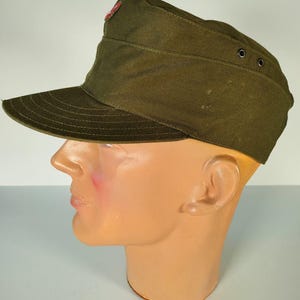 May include: A green military-style cap with a red star emblem on the front. The cap has a visor and a button on the side.