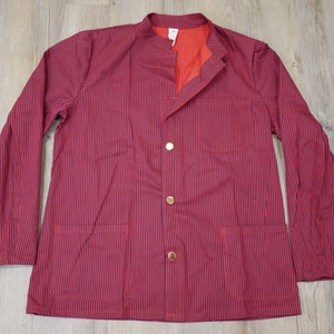 Op de afbeelding: Een rood-blauw gestreept werkjasje met een mandarijnkraag en drie gouden knopen. Het jasje heeft twee zakken aan de voorkant en één zak op de linkermouw.