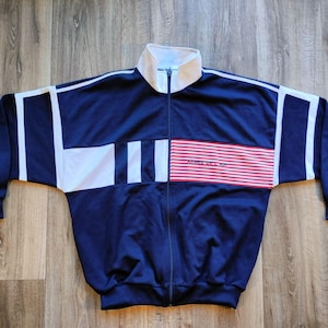 Peut inclure: Veste de survêtement zippée bleu marine avec des accents blancs et rouges. La veste a un col blanc et des rayures blanches sur les manches et la poitrine. La poitrine présente une section rayée rouge et blanche avec du texte.