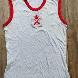 Puede incluir: Camiseta sin mangas blanca con ribetes rojos en el cuello y las sisas. Un emblema bordado en rojo con una corona y remos cruzados está en el pecho. La prenda está hecha de un material suave, probablemente algodón.