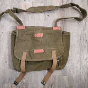 Puede incluir: Un bolso de mensajero de lona verde con correas y hebillas de cuero marrón. El bolso tiene cierre de solapa y una sola correa para el hombro.