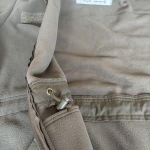 May include: A close-up of the inside of a green fleece jacket. The jacket has a zipper and a pocket with a drawstring. The care label is visible and reads "100% Polyester, Machine Wash, Tumble Dry Low, Do Not Bleach, Do Not Iron".