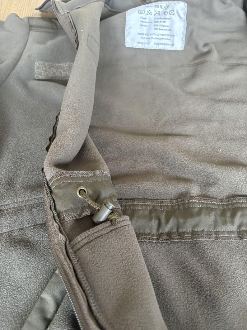 May include: A close-up of the inside of a green fleece jacket. The jacket has a zipper and a pocket with a drawstring. The care label is visible and reads "100% Polyester, Machine Wash, Tumble Dry Low, Do Not Bleach, Do Not Iron".