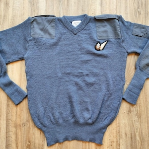 Peut inclure: Un pull bleu à col en V avec des patchs renforcés sur les épaules et les coudes. Le pull présente un petit écusson brodé sur la poitrine. Le vêtement est en maille.