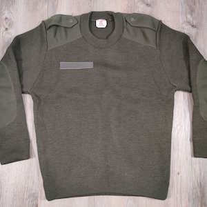 Op de afbeelding: Een groene militaire trui met elleboogpatches en een klein patch op de borst. De trui heeft een ronde hals en lange mouwen.