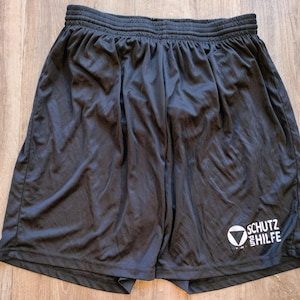 Puede incluir: Pantalones cortos deportivos negros con cintura elástica. Los pantalones cortos tienen un logotipo blanco que dice "SCHUTZ HILFE". Hechos de un material ligero y ligeramente brillante.