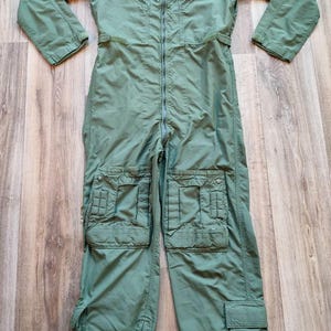 Peut inclure: Combinaison de vol vert olive avec plusieurs poches sur les jambes et la poitrine. Le vêtement une pièce a une fermeture éclair pleine longueur, des écussons sur les épaules et un col. Les manches ont des poches zippées et les jambes ont des fermetures éclair aux chevilles.