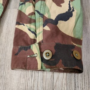 May include: Close-up of the cuff of a camouflage jacket with two brown buttons. The fabric is a brown, green, and black camouflage pattern.