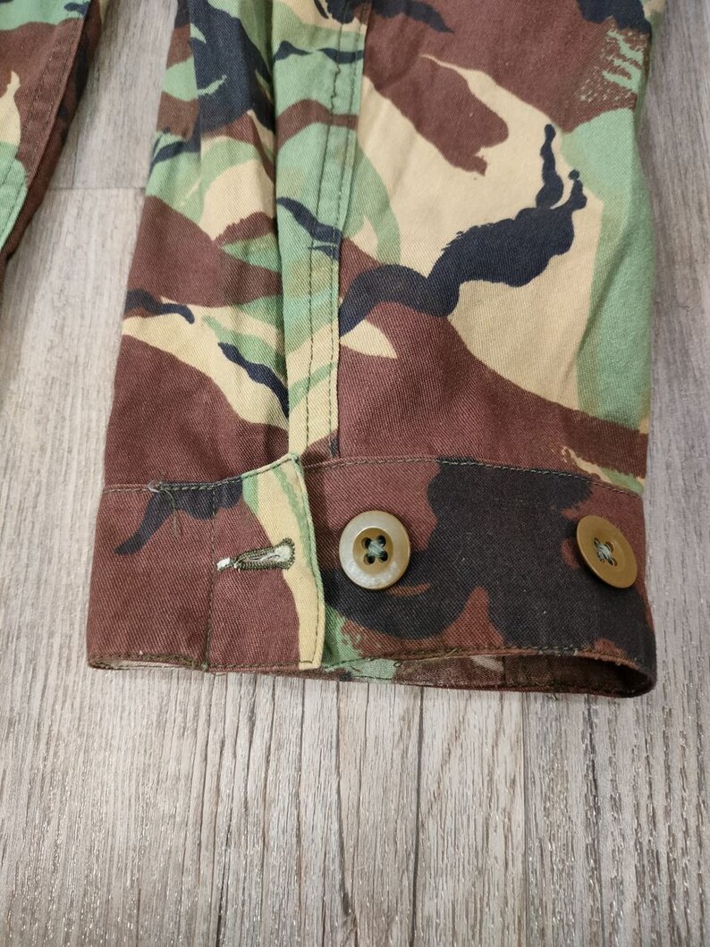 May include: Close-up of the cuff of a camouflage jacket with two brown buttons. The fabric is a brown, green, and black camouflage pattern.
