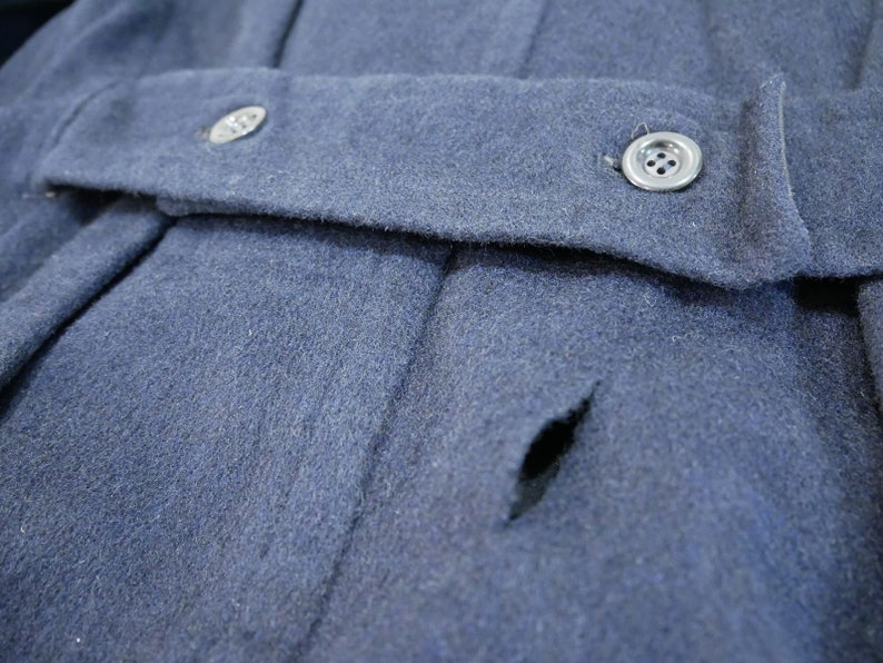 May include: A close-up of a dark blue wool jacket with a small tear in the fabric. The jacket has a button closure and a single button on the front.