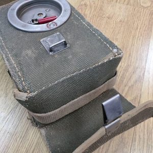 May include: A green canvas bag with a metal latch and a round metal compartment with a red lever. The bag has a strap and a metal clip.