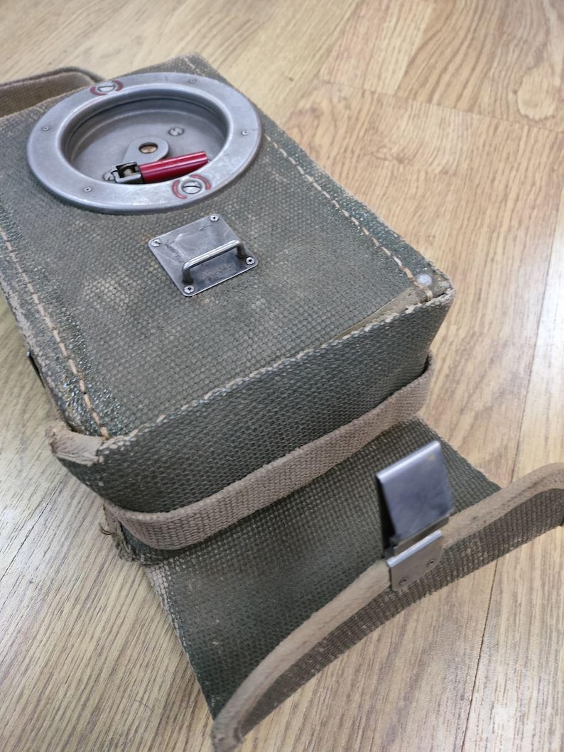 May include: A green canvas bag with a metal latch and a round metal compartment with a red lever. The bag has a strap and a metal clip.