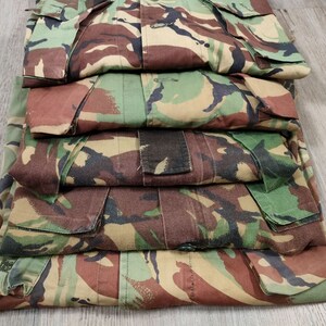 May include: A stack of five camouflage-patterned shirts in shades of green, brown, and tan. The shirts are folded neatly and have a button-up closure.