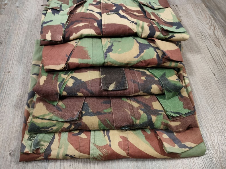 May include: A stack of five camouflage-patterned shirts in shades of green, brown, and tan. The shirts are folded neatly and have a button-up closure.