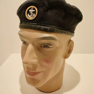 Op de afbeelding: Een zwarte baret met een gouden ankerembleem op de voorkant. De baret wordt gedragen op een mannequinhoofd.