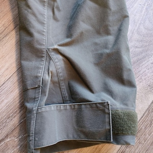 May include: Close-up of a section of olive green fabric, possibly a jacket or pants. The image shows a cuff with a Velcro closure and detailed stitching. The fabric has a slightly worn appearance.