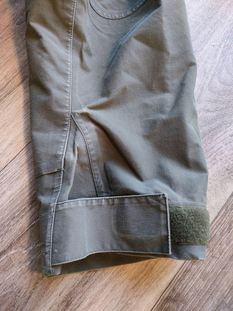 May include: Close-up of a section of olive green fabric, possibly a jacket or pants. The image shows a cuff with a Velcro closure and detailed stitching. The fabric has a slightly worn appearance.