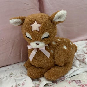 May include: A plush deer toy with brown textured fur, white spots on its back, and a light pink star patch on its forehead. The deer has closed eyes with stitched eyelashes, a small white muzzle, and a light pink satin ribbon tied around its neck.