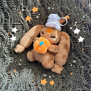 Könnte beinhalten: Ein brauner Plüsch-Hasen-Schlüsselanhänger mit langen Ohren, einer weißen Mütze im Stil von Schlagsahne und einem orangefarbenen Kürbis in den Pfoten. Der Hase hat einen kleinen orangefarbenen Stern auf der Stirn und einen silbernen Metallring.