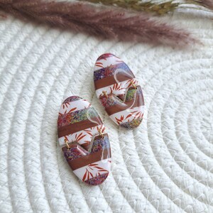May include: A pair of oval-shaped earrings with a white and brown striped pattern. The earrings have a glittery finish and feature a red and white floral design.