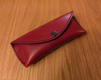 May include: A red leather glasses case with a black stitched flap and a silver button closure.