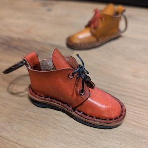 Peut inclure: Deux porte-clés en forme de bottines en cuir. L'un est d'un rouge orangé vif, l'autre d'un bronzage plus clair. Les deux ont des lacets et un anneau porte-clés. Les bottes sont détaillées avec des coutures et un bout arrondi.