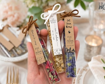 May include: Three clear glass test tubes filled with dried tea leaves and flower petals. Each tube is sealed with a cork and features a personalized paper tag tied with a brown or white string. The tags display text including "The Perfect Blend," "Let Love Brew," and "Thank you for coming!" along with the names "Eve & Adam" and the date "04/12/2024."