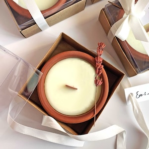 May include: A set of small terracotta candle holders with a white candle inside each. The candle holders are packaged in clear plastic boxes with brown lids and tied with white ribbon. A white tag with the text "Eve & Adam" is attached to the ribbon.