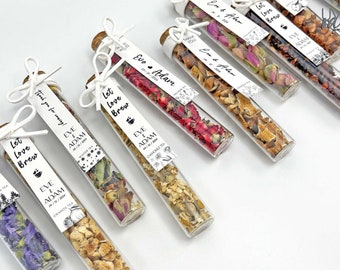 May include: An assortment of small glass tubes filled with dried flowers and herbs, each with a white label that reads "Let Love Brew" and "Eve & Adam" with a wedding date. The labels also include the type of tea, such as "Orange Tea" and "Chamomile Tea".