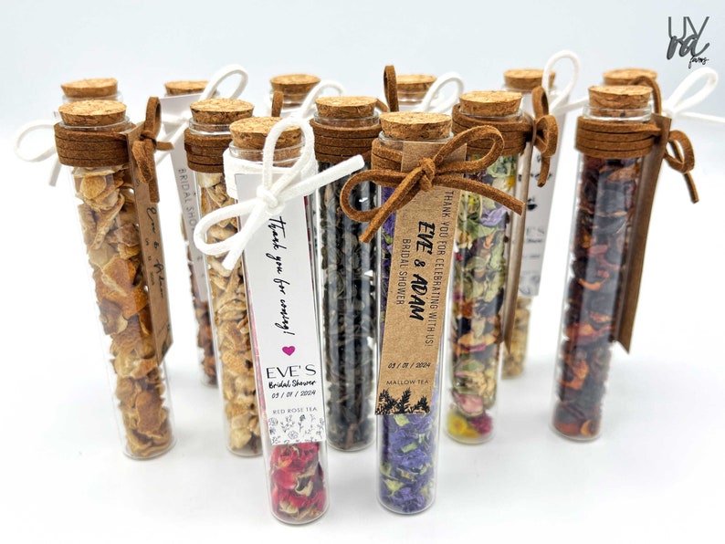 May include: A collection of small glass tubes filled with dried flowers, herbs, and fruit. Each tube is tied with a brown ribbon and a small tag with the text "Thank you for celebrating with us Eve & Adam Bridal Shower" and "Thank you for coming! Eve's Bridal Shower 05/11/2024 Red Rose Tea".